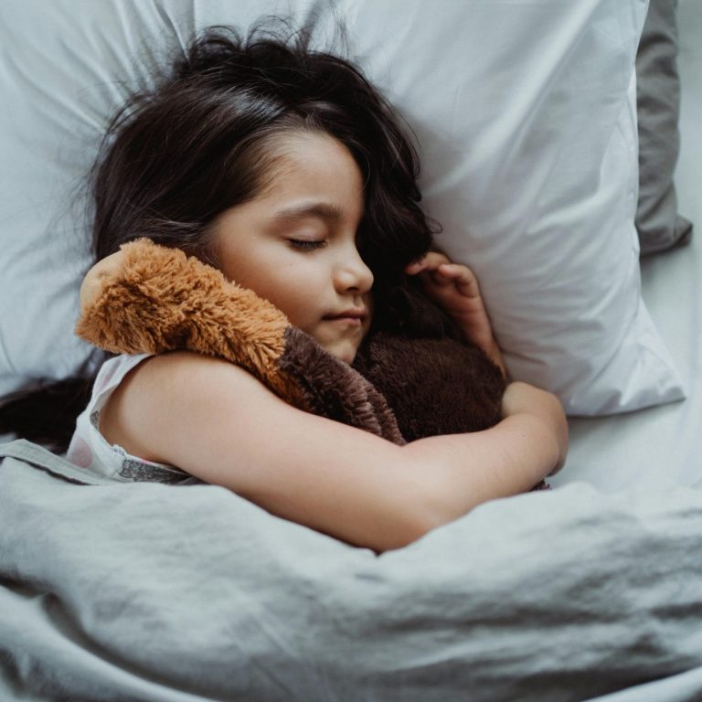 Een peutermeisje slaapt met een knuffel in haar armen, ontspannen op een bed.