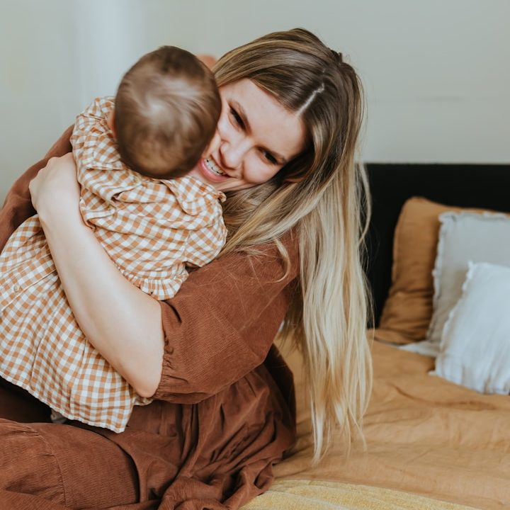 Een vrouw omarmt een baby op een bed met een beige en witte dekbed.