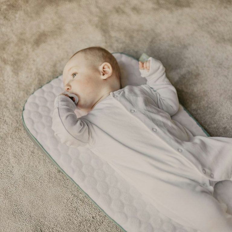 Een baby ligt op een speelkleed en steekt een vinger in de mond.