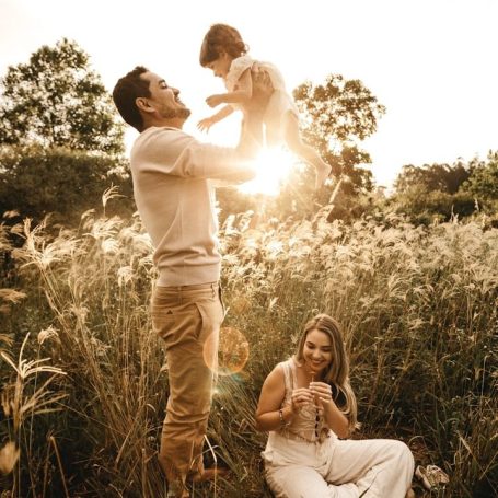 Vader tilt kind op in de lucht, terwijl moeder op de grond met bloemen speelt.